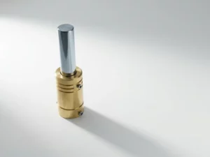 Brass hydraulic cylinder with chrome piston rod extending upward on white surface, minimalist composition with geometric shadows