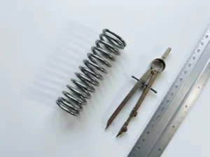 Titanium spring mechanism with drafting compass and ruler on white surface casting geometric shadows in minimalist workspace
