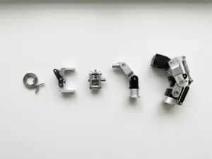 Five precision-machined metal prototypes arranged by complexity on white laboratory workbench, from simple spring to intricate exoskeleton joint assembly