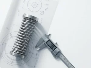 Precision-engineered spring mechanism with aligned coils on white technical drawing table, digital caliper and blueprints nearby