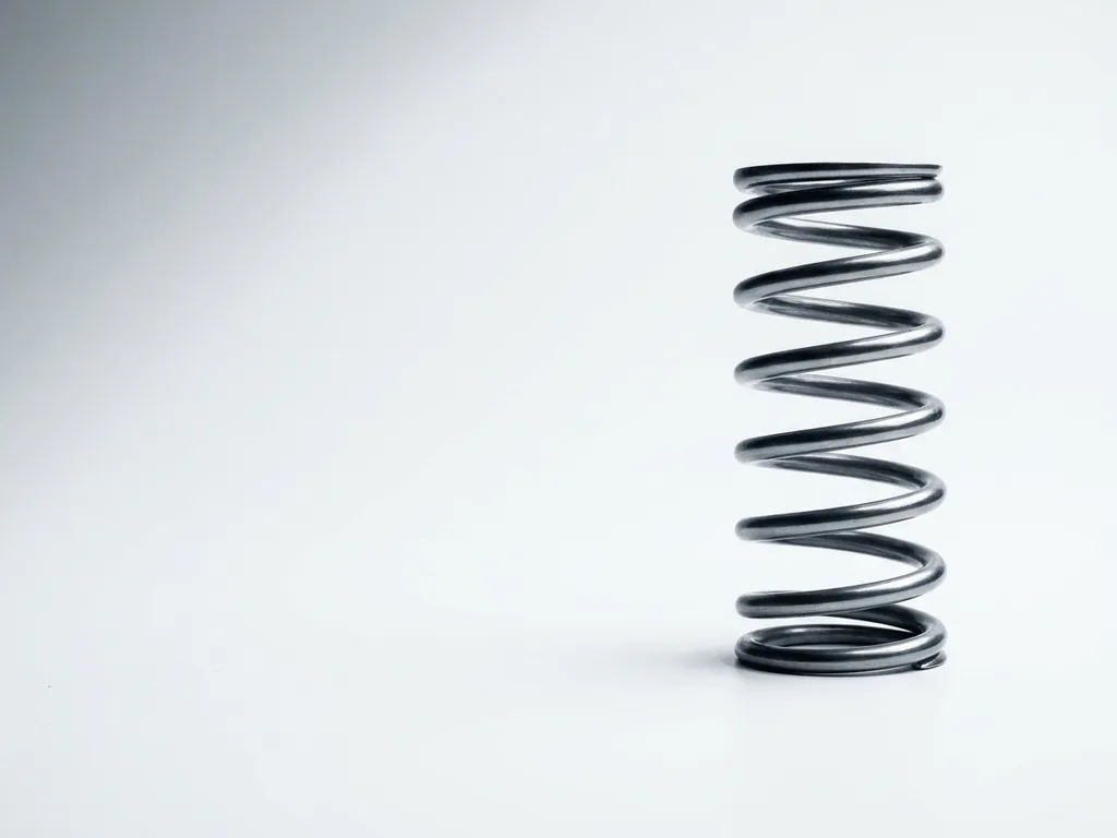Polished steel compression spring standing vertically on white surface with dramatic lighting creating silver to charcoal gradients